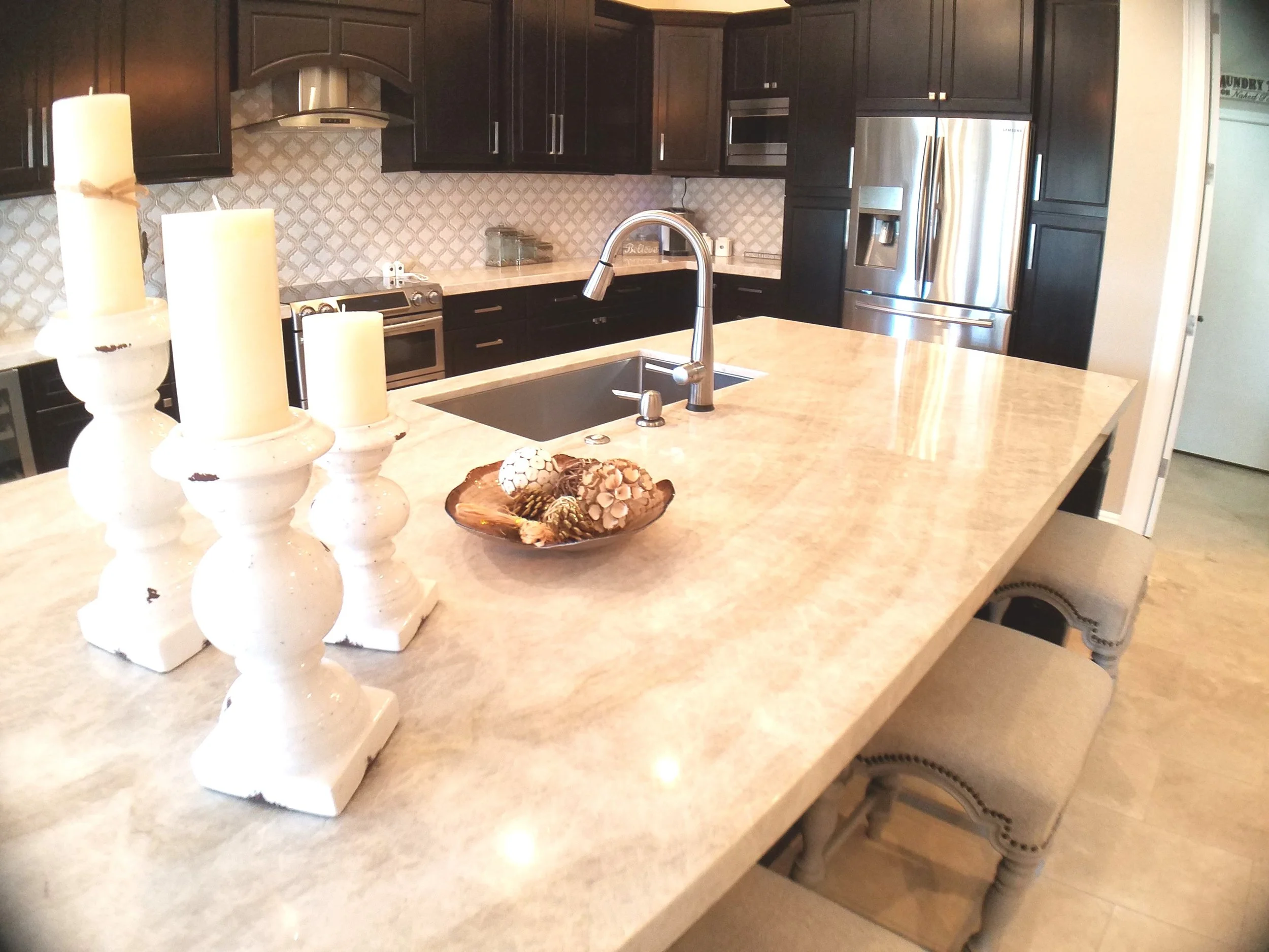 Espresso brown kitchen cabinets with a large pale quartzite island, arabesque tile backsplash and stainless appliances
